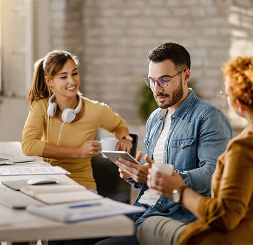 Three colleagues sitting around a table with coffee, looking at a tablet together during a marketing planning session
