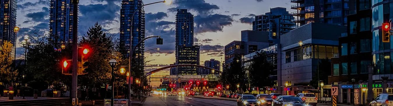 Surrey City Centre at dusk, looking down a lit boulevard of traffic towards the residential towers and the arched SkyTrain guideway.