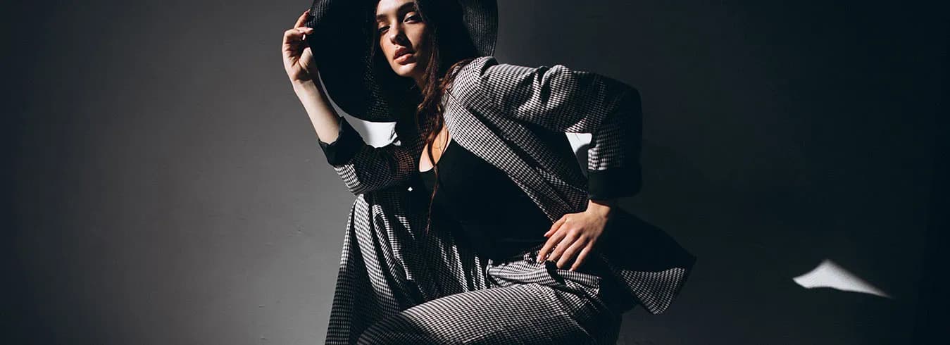 A model in a black-and-white patterned coat and trousers seated against a dark studio backdrop, one arm raised above her head.