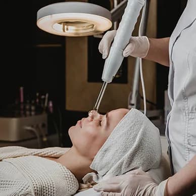 A client reclining under a treatment lamp while a gloved practitioner performs a facial procedure.