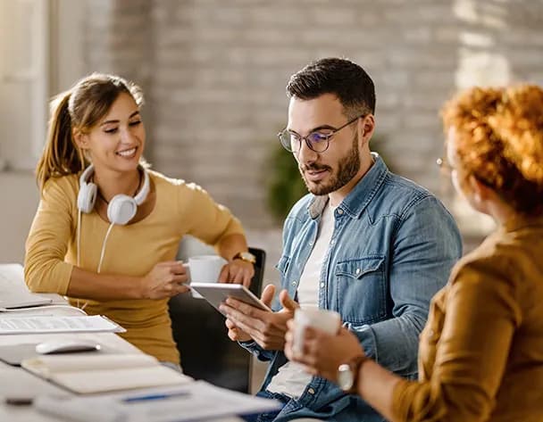 Three colleagues around a table in a bright office, one showing a tablet to the others over coffee.