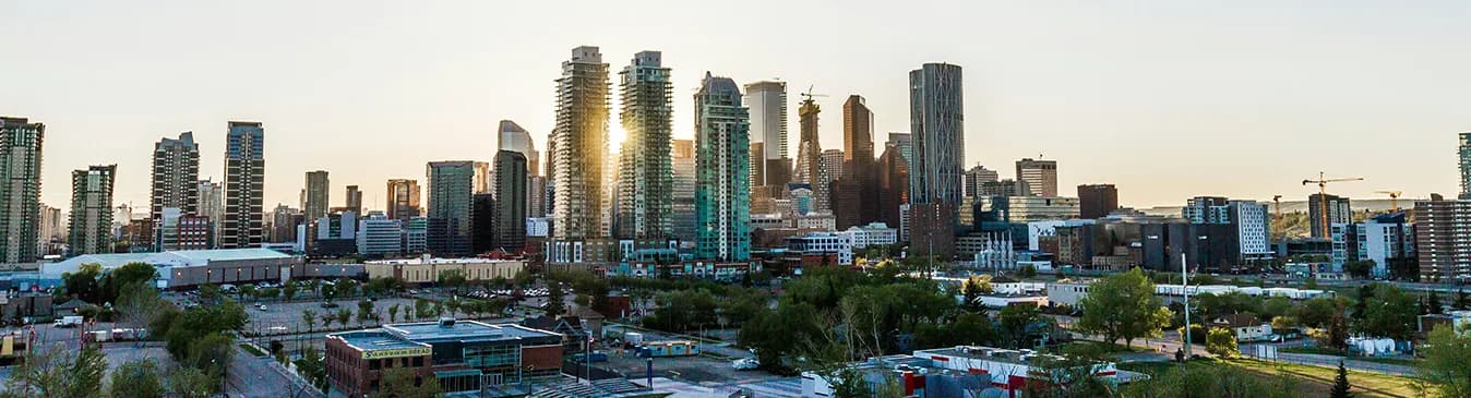 The downtown Calgary skyline photographed at sunset, the low sun flaring between the glass office towers, with parkland and low-rise blocks in the foreground.