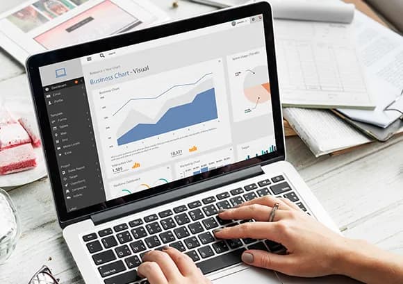 Hands typing on a laptop that displays a “Business Chart – Visual” marketing dashboard, on a white desk scattered with printed reports and charts.