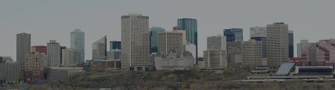 The downtown Edmonton skyline on an overcast autumn day, with the château-roofed Hotel Macdonald in front of the office towers and the river valley treeline below.
