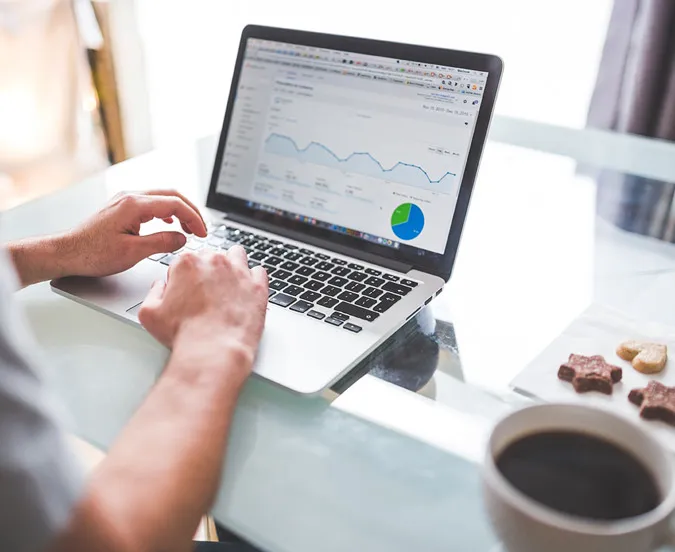 A person typing on a laptop showing a web analytics dashboard with a traffic line graph and a pie chart, beside a cup of black coffee on a glass table.