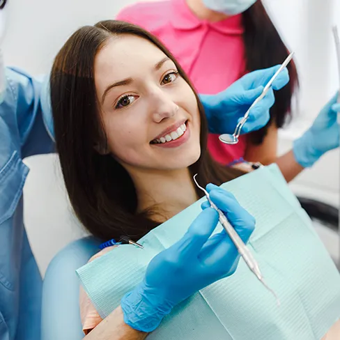 A smiling patient in a dental chair while gloved hands hold a mirror and scaler above her.