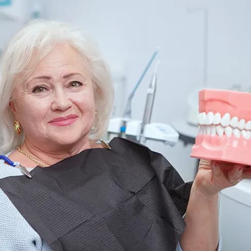 An older woman in a dental chair smiling and holding up an oversized model of a set of dentures.