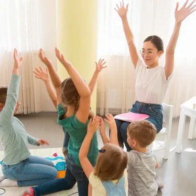 A daycare teacher and a group of young children sitting on the floor of a bright playroom with their arms stretched overhead.