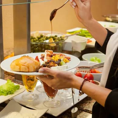 A hand drizzling sauce from a spoon over a plated dish at a restaurant buffet counter.