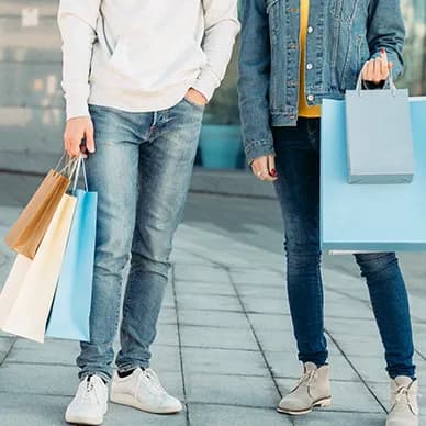 Two shoppers photographed from the waist down on a city sidewalk, each carrying paper shopping bags.