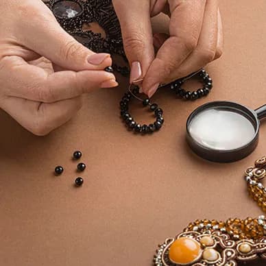 Close-up of two hands threading black beads into a hoop earring, beside a jeweller’s magnifying loupe.